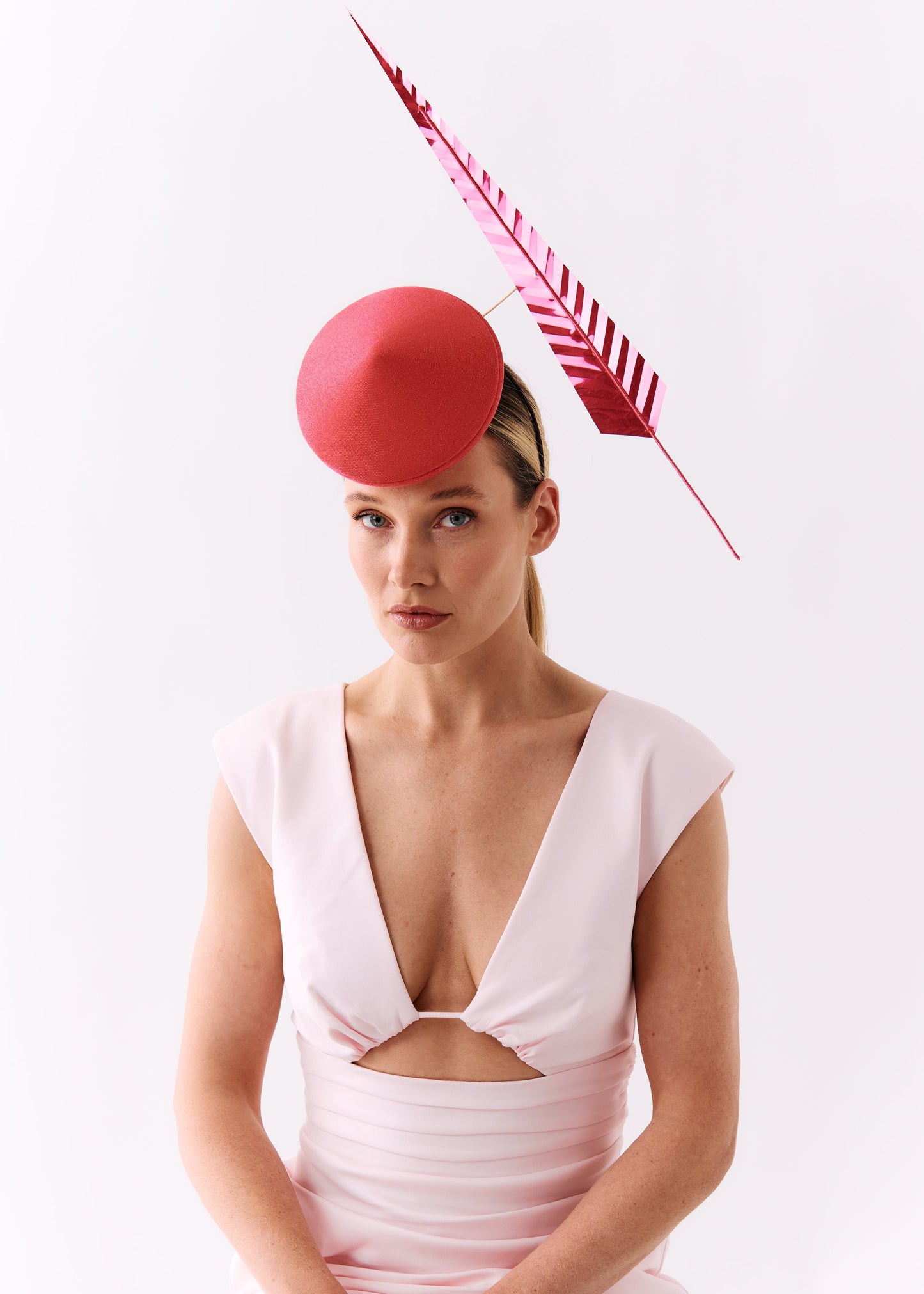 A woman wearing a pale pink dress and a bright pink fascinator hat with a matching feather, posed against a solid white studio background.