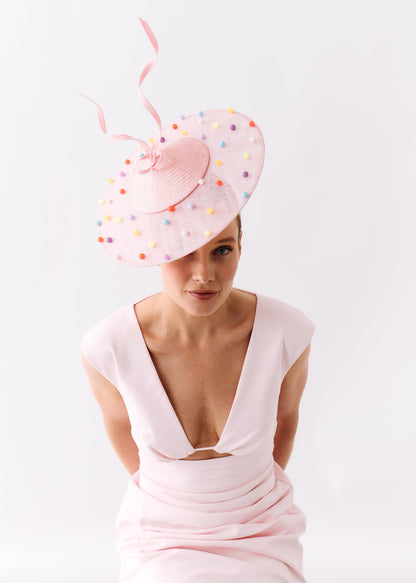 A woman wearing a pale pink dress and a matching wide-brimmed pink fascinator hat decorated with small multicolored beads, posed against a clean white studio background.