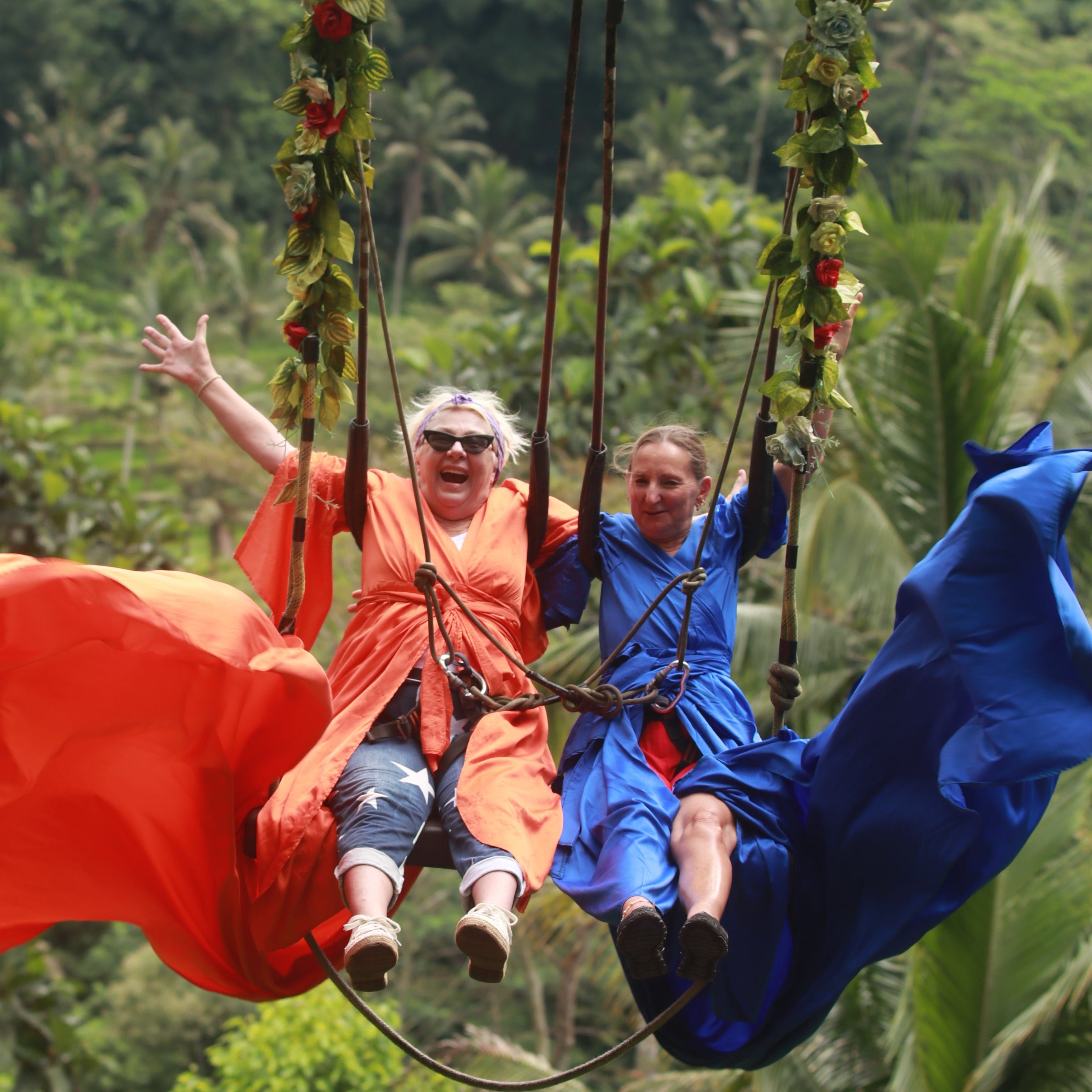 Two people in colorful costumes on a big swing outdoors with greenery in the background.