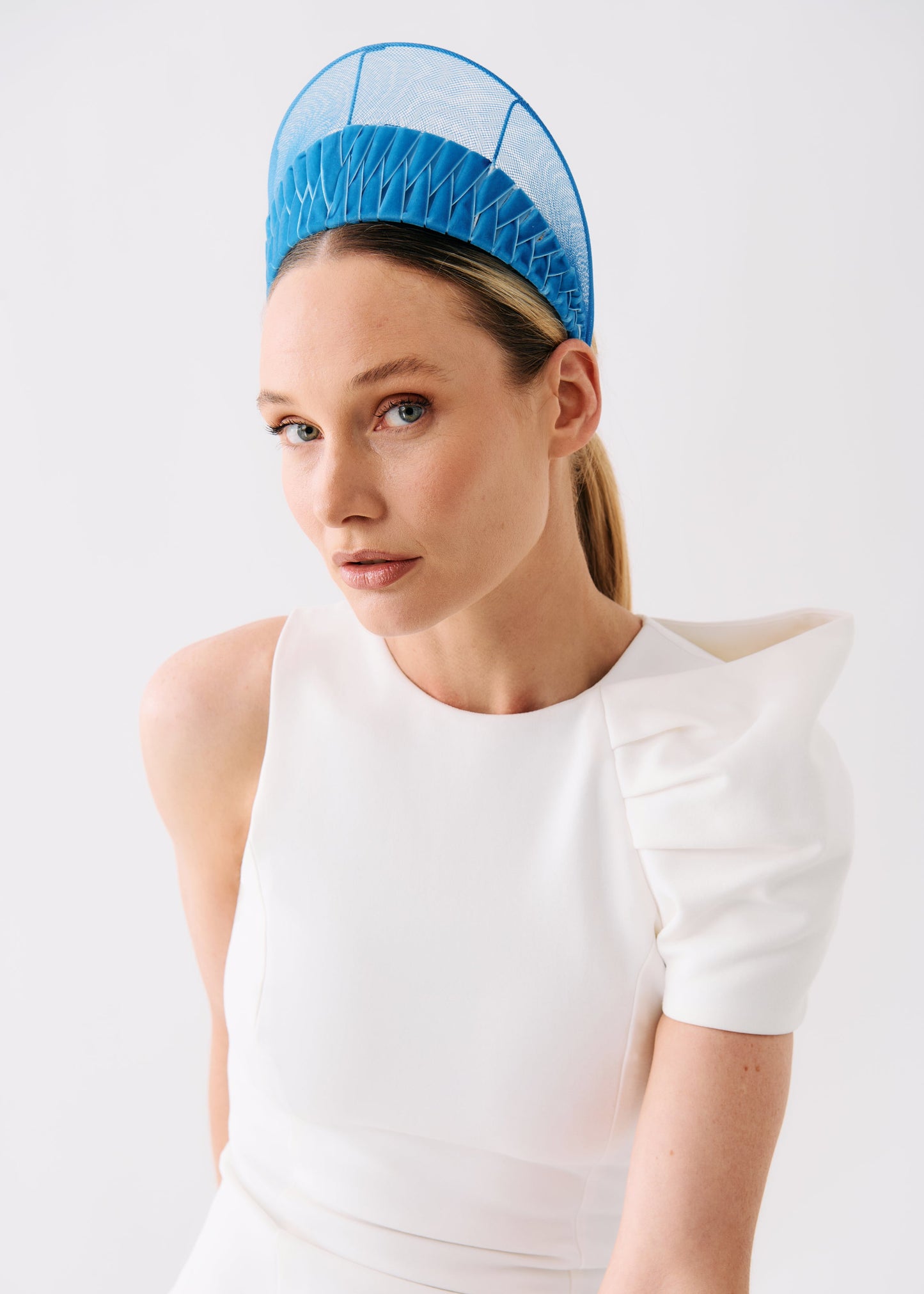 A woman wearing a bright blue structured fascinator headpiece and a white sleeveless dress with puffed shoulders, photographed against a clean light gray studio background.