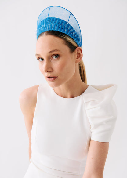 A woman wearing a bright blue structured fascinator headpiece and a white sleeveless dress with puffed shoulders, photographed against a clean light gray studio background.