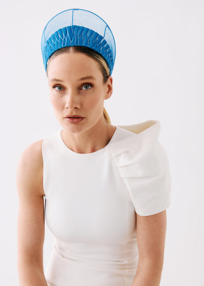 A woman wearing a structured white sleeveless dress with a dramatic ruffled shoulder detail and a bright blue pleated headpiece, photographed against a clean white studio background.