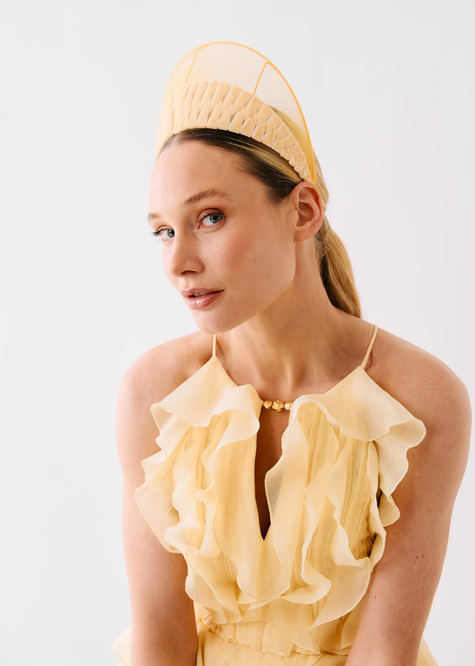 A woman wearing a pale yellow ruffled halter dress and a matching structured fascinator headpiece, photographed against a clean white studio background.