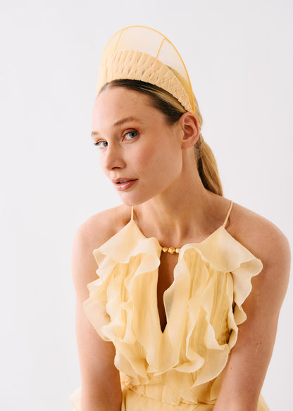 A woman wearing a pale yellow ruffled halter dress and a matching structured fascinator headpiece, photographed against a clean white studio background.
