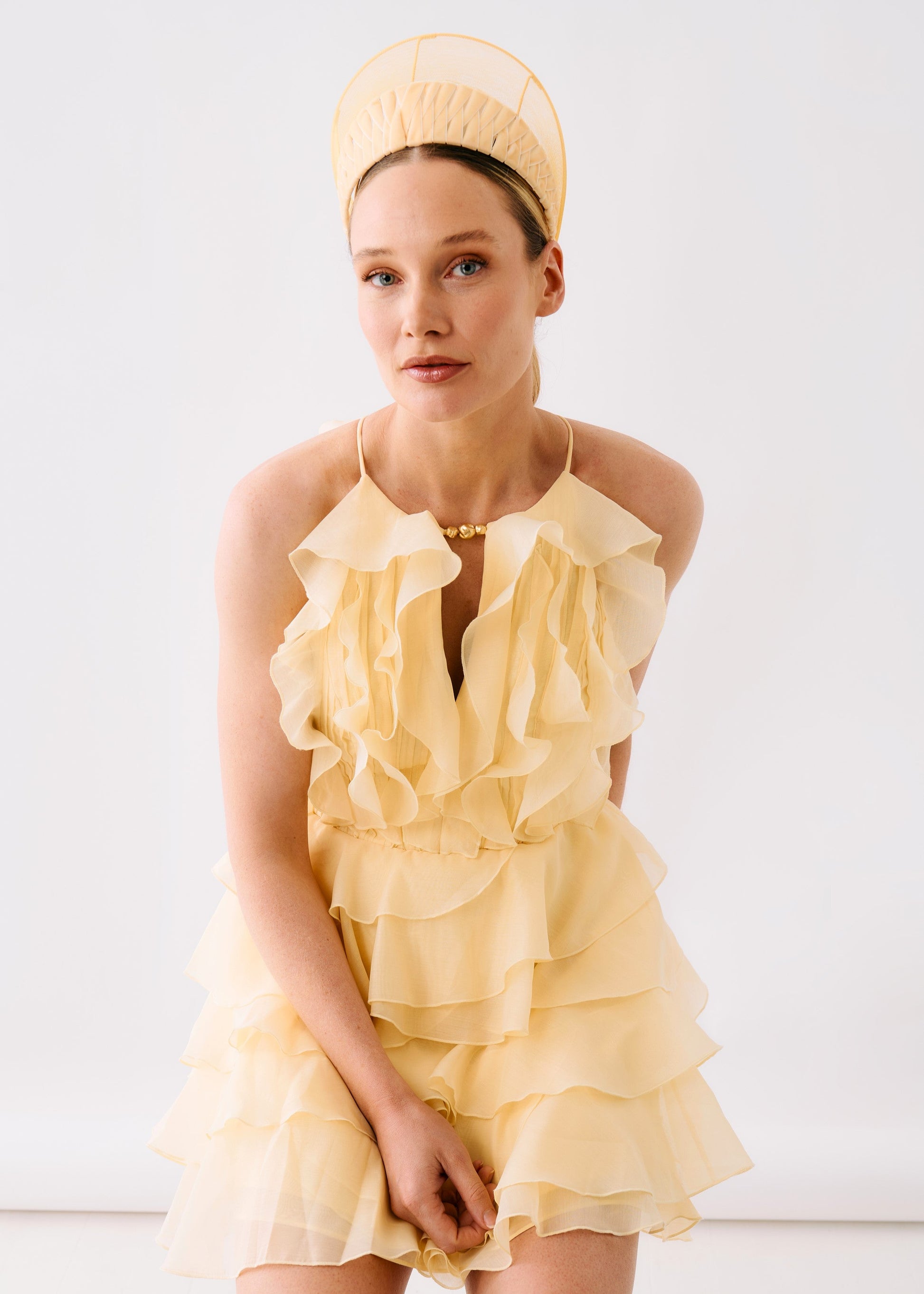 A model wearing a pale yellow ruffled halter dress with a matching sheer headband, posed against a clean white studio background.