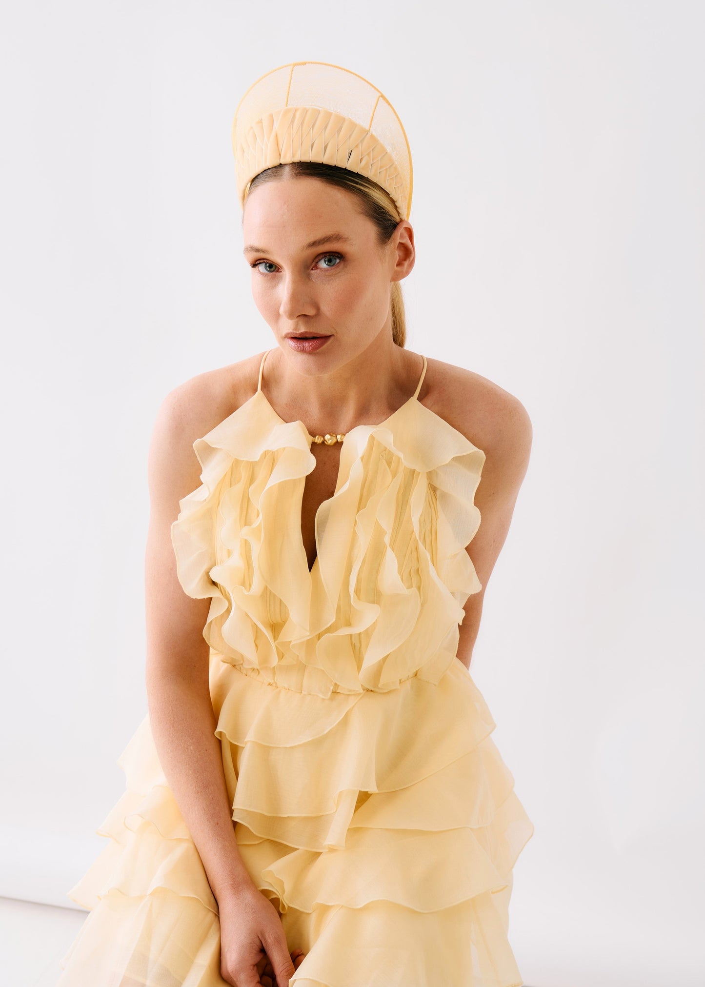 A model wearing a pale yellow ruffled halter dress with a matching structured headpiece, posed against a clean white studio background.