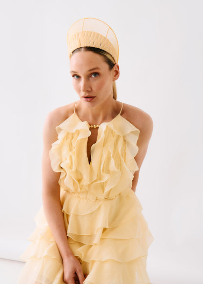 A model wearing a pale yellow ruffled halter dress with a matching structured headpiece, posed against a clean white studio background.