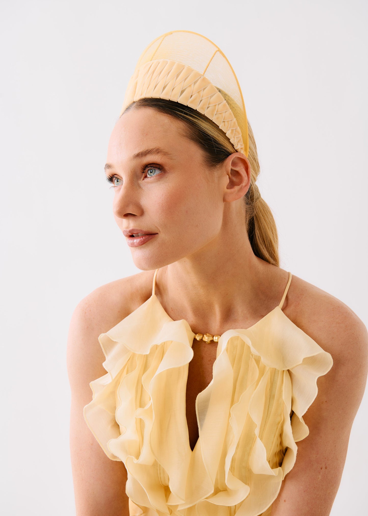 A woman wearing a pale yellow ruffled halter dress and a matching structured fascinator headpiece, photographed against a clean light gray studio background.