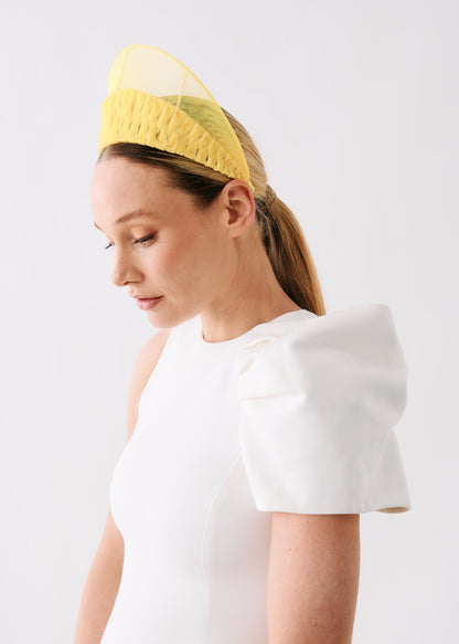 A woman wearing a white dress with dramatic puffed shoulders and a yellow woven fascinator headpiece, photographed against a clean light gray studio background.