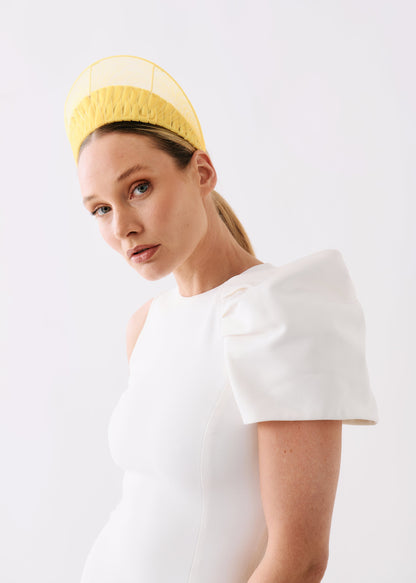 A woman wearing a structured white dress with dramatic puffed shoulders and a yellow mesh headband, photographed against a plain light gray studio background.
