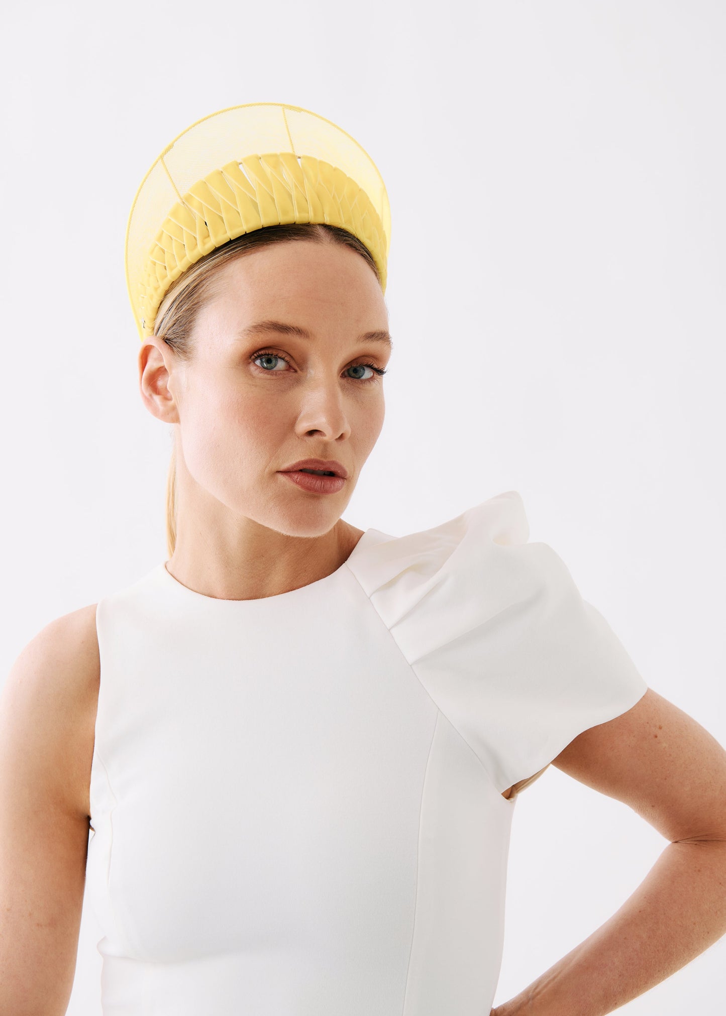 A woman wearing a white sleeveless dress with a dramatic ruffled shoulder detail and a yellow structured headpiece, photographed against a clean white studio background.